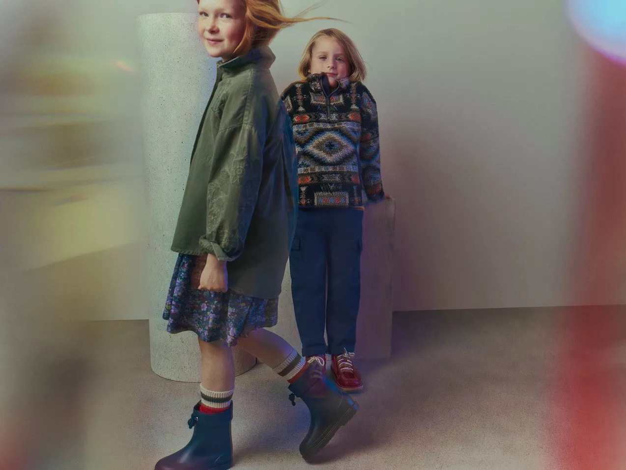 Girl in army green denim shirt and floral skirt, walking in front of boy in patterned sweater and denim trousers. 