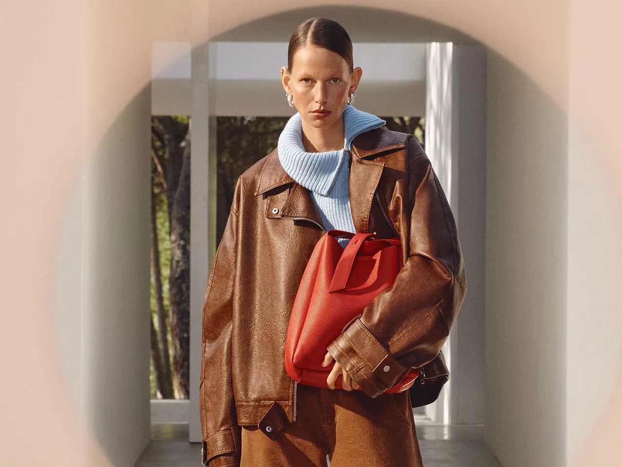A woman in a brown leather jacket and blue sweater holding a red handbag