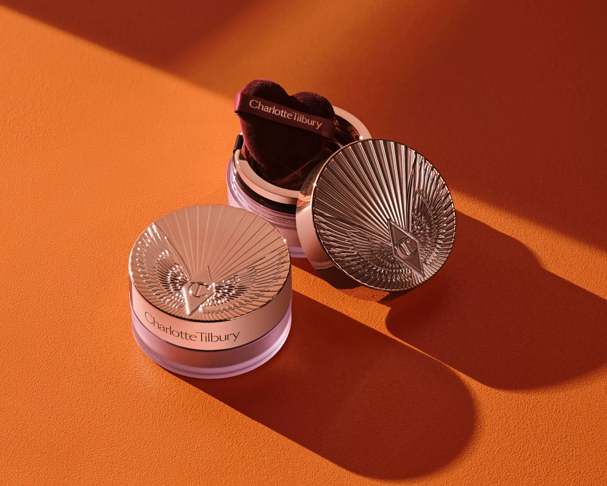 Two Charlotte Tilbury setting powders, with a heart-shaped powder puff on a flat orange-coloured backdrop