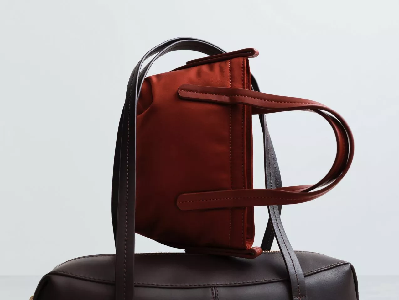 Red mini tote handbag with long leather shoulder strap, balancing on a brown leather bag