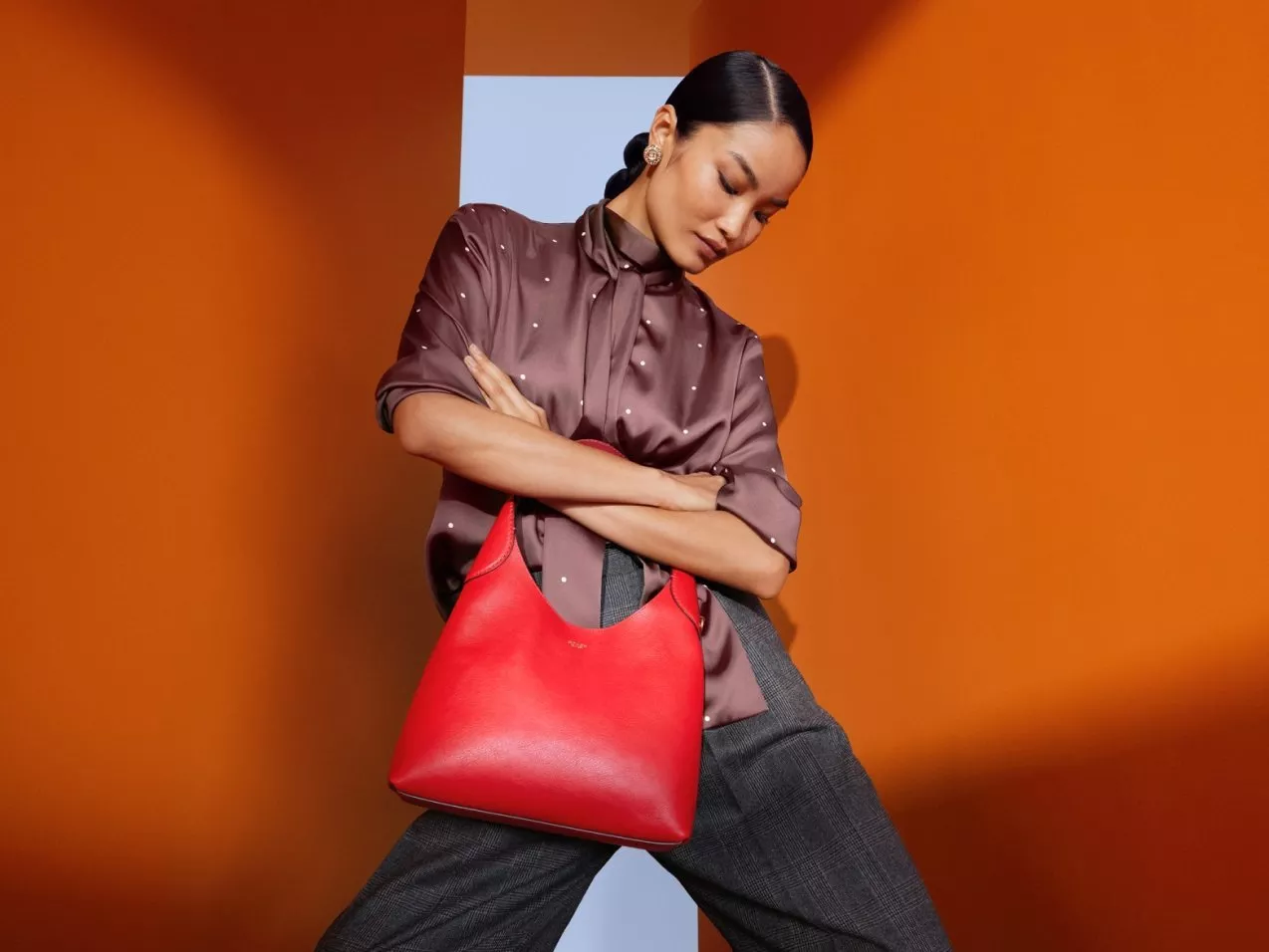 Fashion model posing with a red leather hobo handbag, wearing a satin blouse and grey trousers on orange backdrop