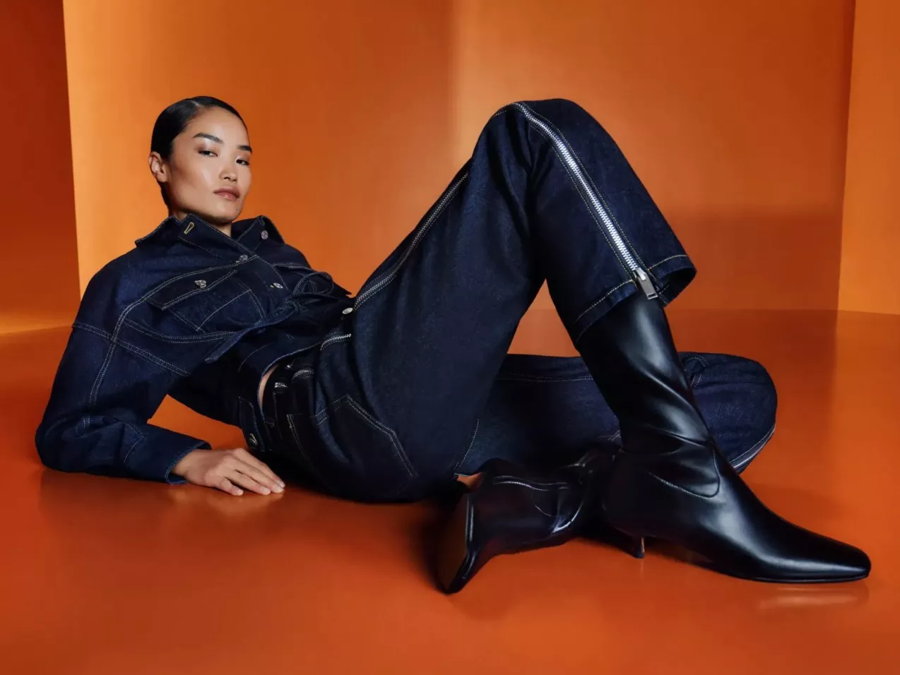A woman in dark denim jumpsuit and black leather knee-high boots posing on an orange studio backdrop