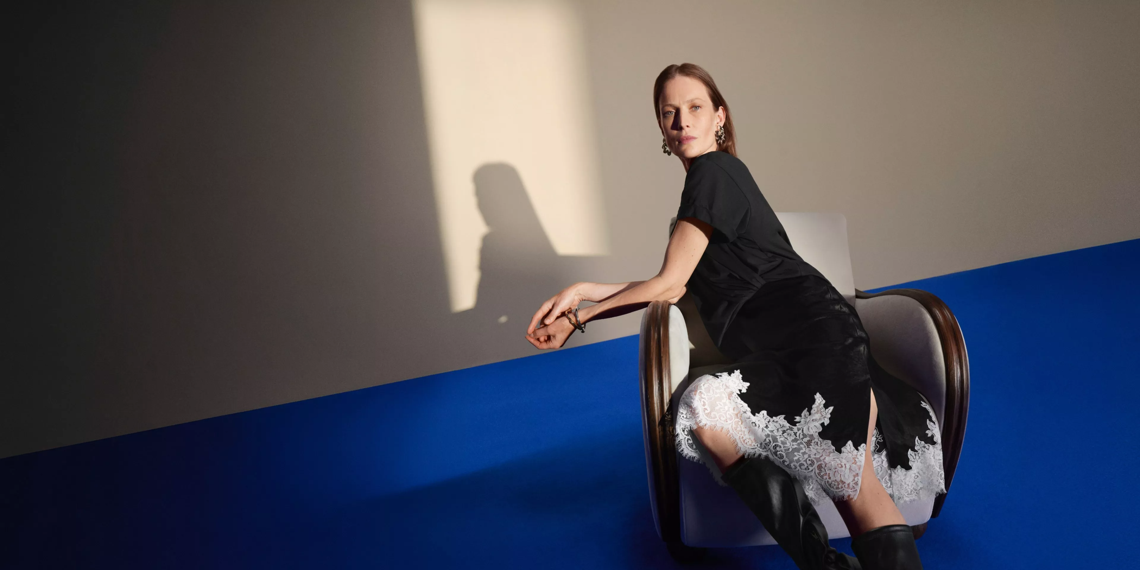 Woman in black dress with lace hem, seated on chair against blue and grey studio wall
