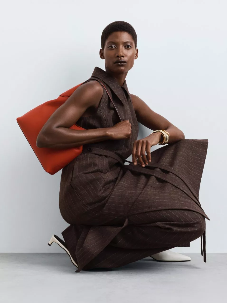 Fashion model crouching in brown pinstripe sleeveless suit, holding orange tote bag, studio portrait on grey backdrop