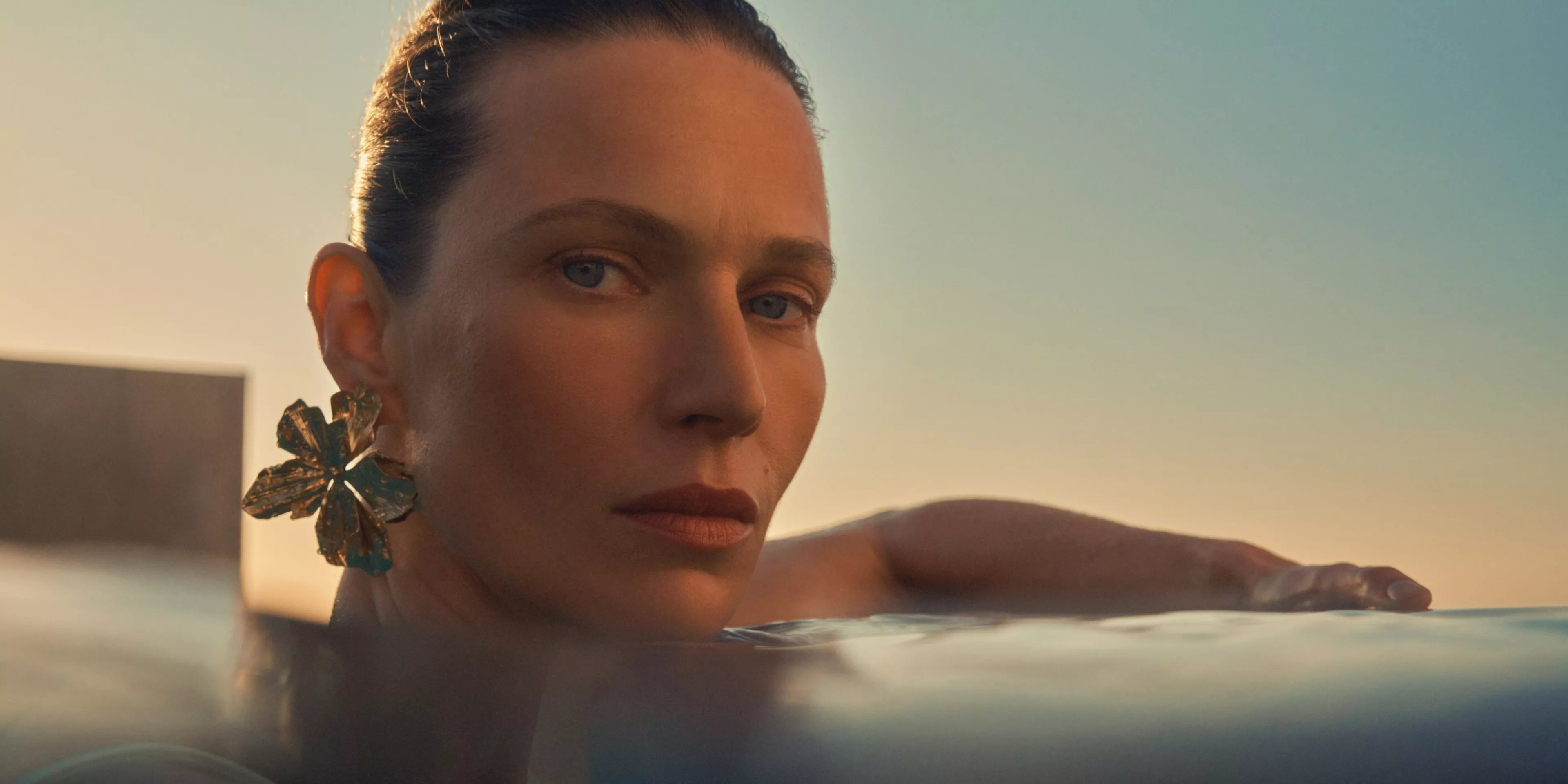 women wearing eye makeup in a swimming pool with water up to her neck with a sunset behind her 