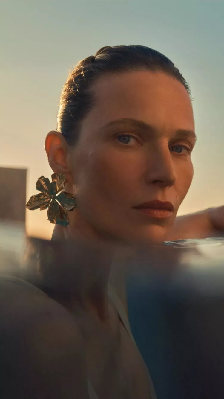 women wearing eye makeup in a swimming pool with water up to her neck with a sunset behind her 