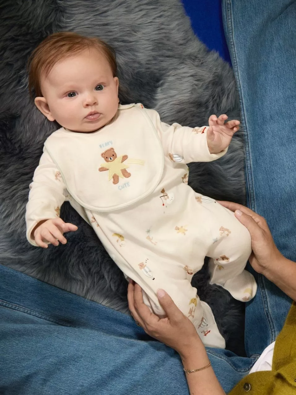 Baby in cream sleepsuit and teddy bib, lying on grey faux fur blanket, held by parent