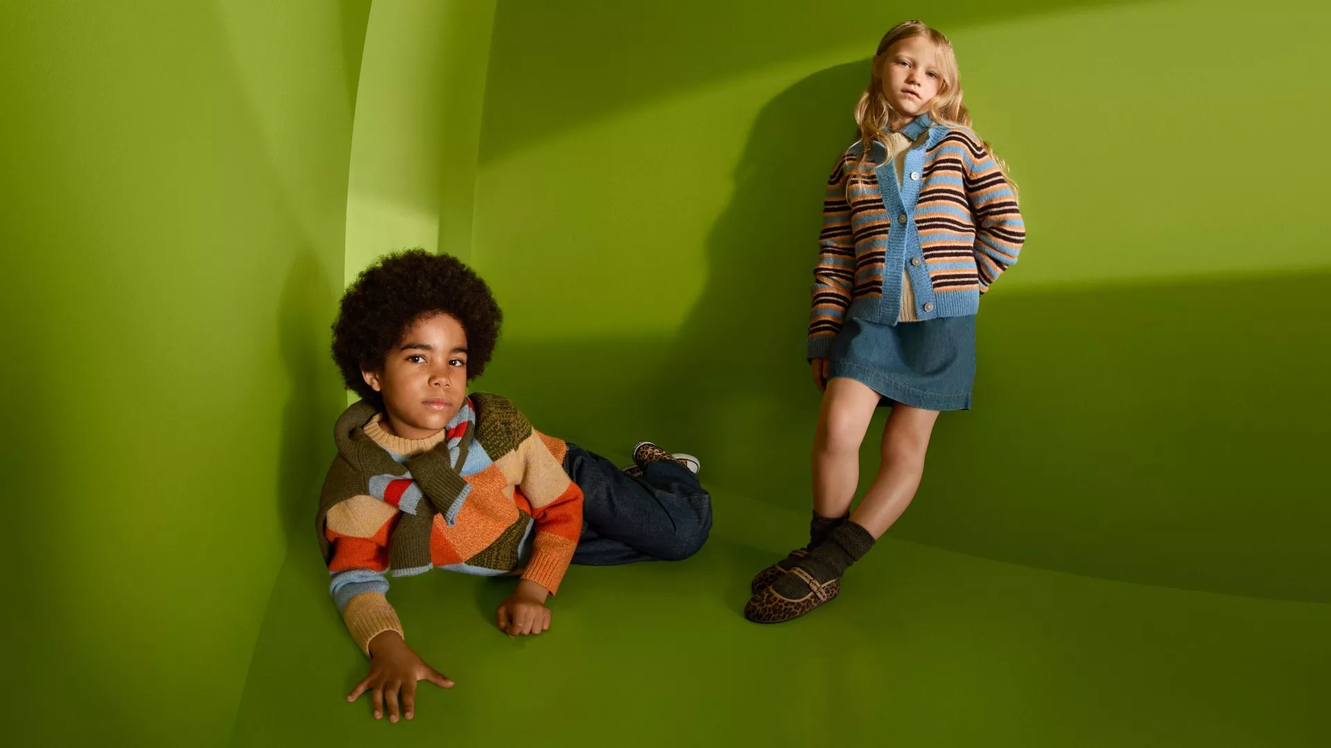 Two children modelling colourful knitwear and denim outfits in a green studio