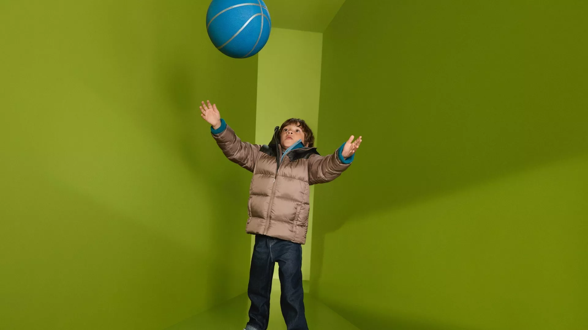 Child in a puffer jacket tossing a blue basketball indoors against a bright green wall