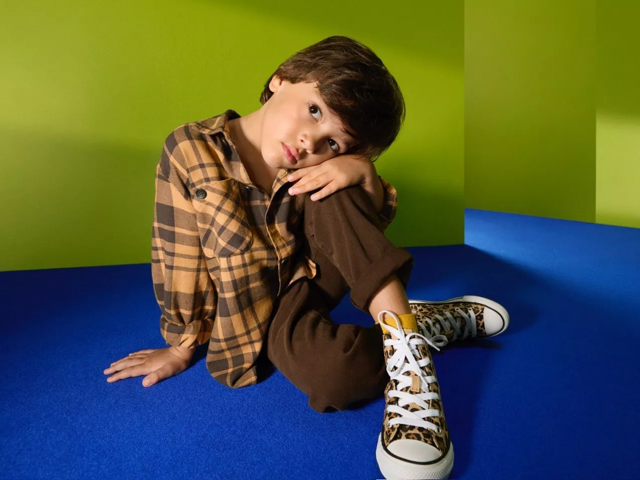 Young boy in checked shirt and brown trousers sitting on blue floor, wearing leopard print trainers on green backdrop