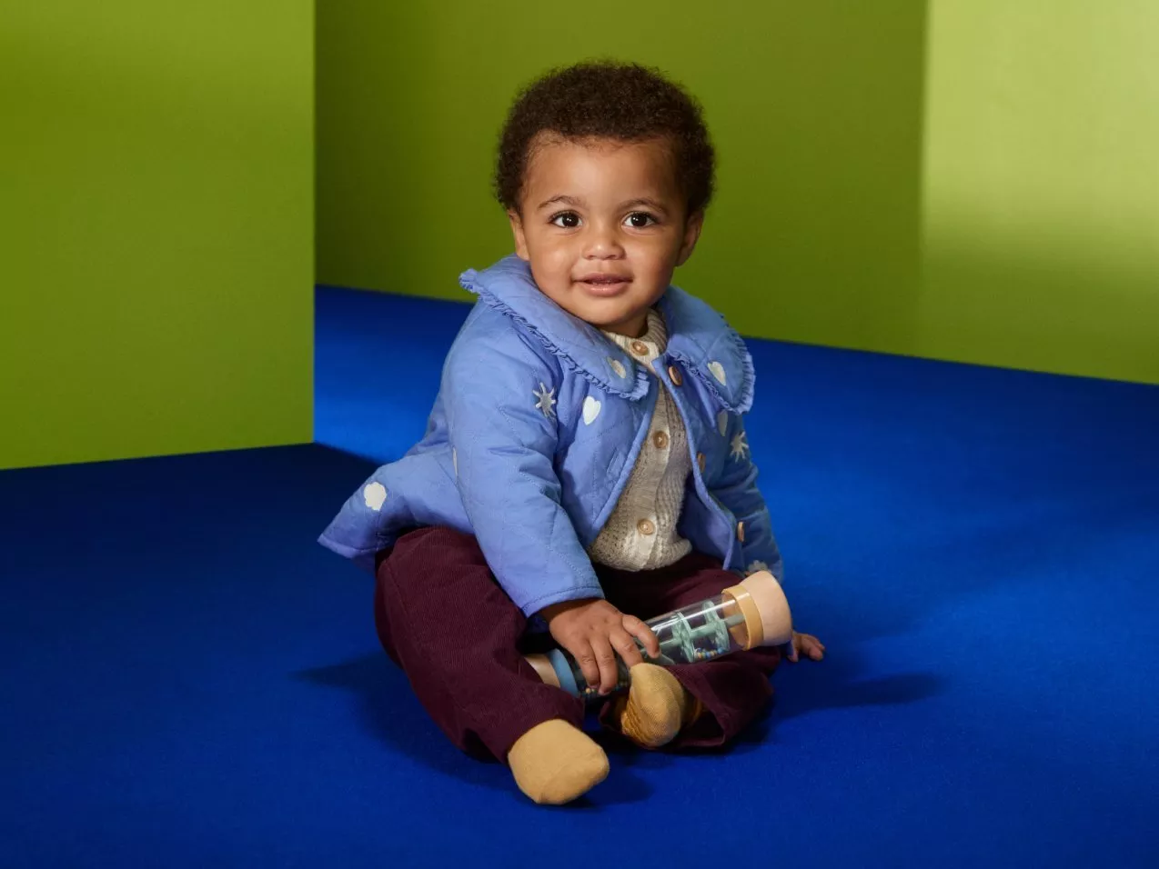 Smiling toddler in a blue padded jacket sitting on a blue sofa, holding a toy bottle, green background.