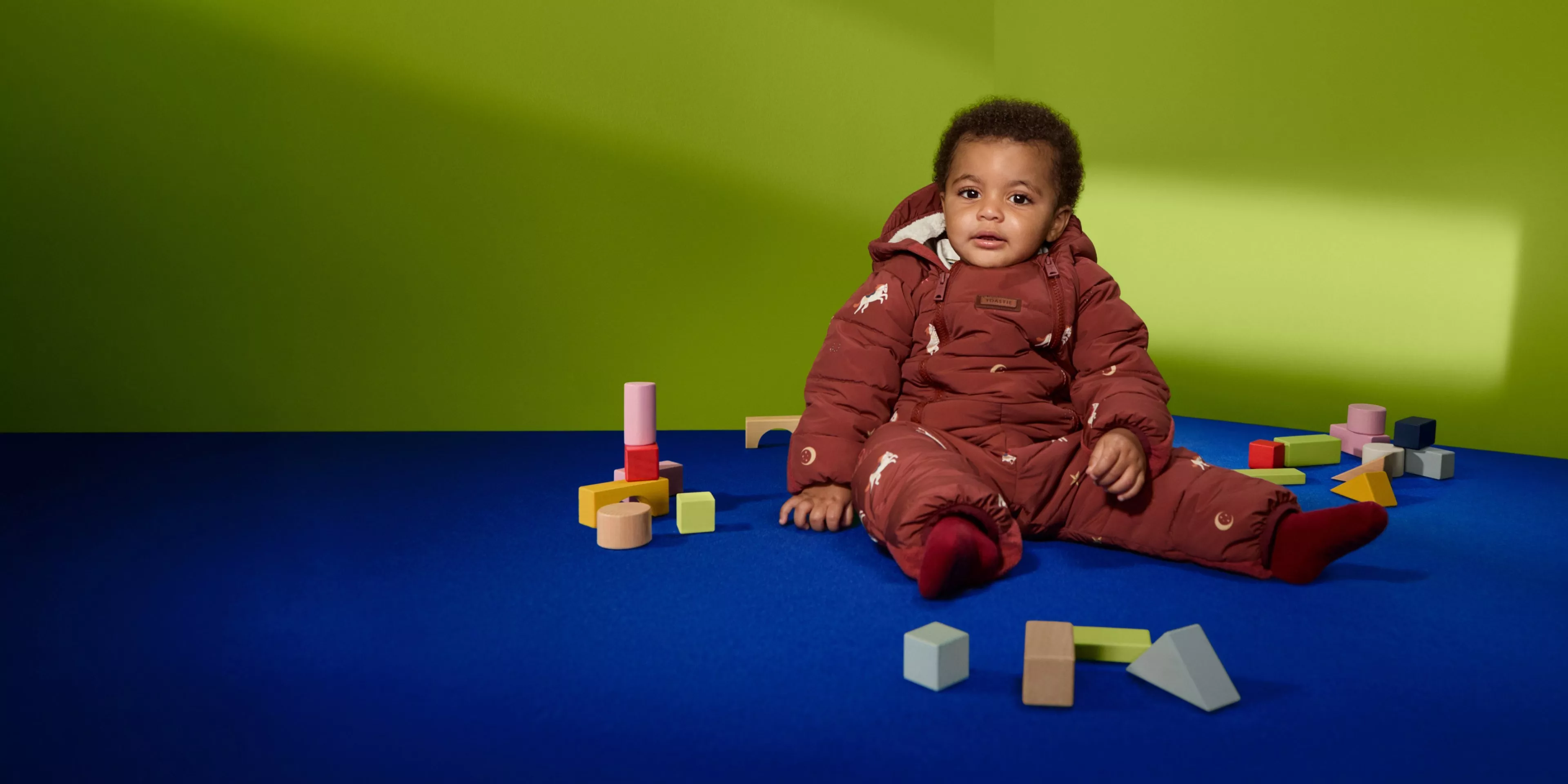 Baby in a maroon padded snowsuit sitting on a blue mat with wooden blocks against a green backdrop