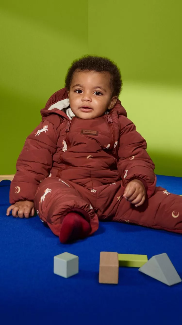 Baby wearing a brown padded snowsuit with hood, sitting on a blue mat against a green studio background