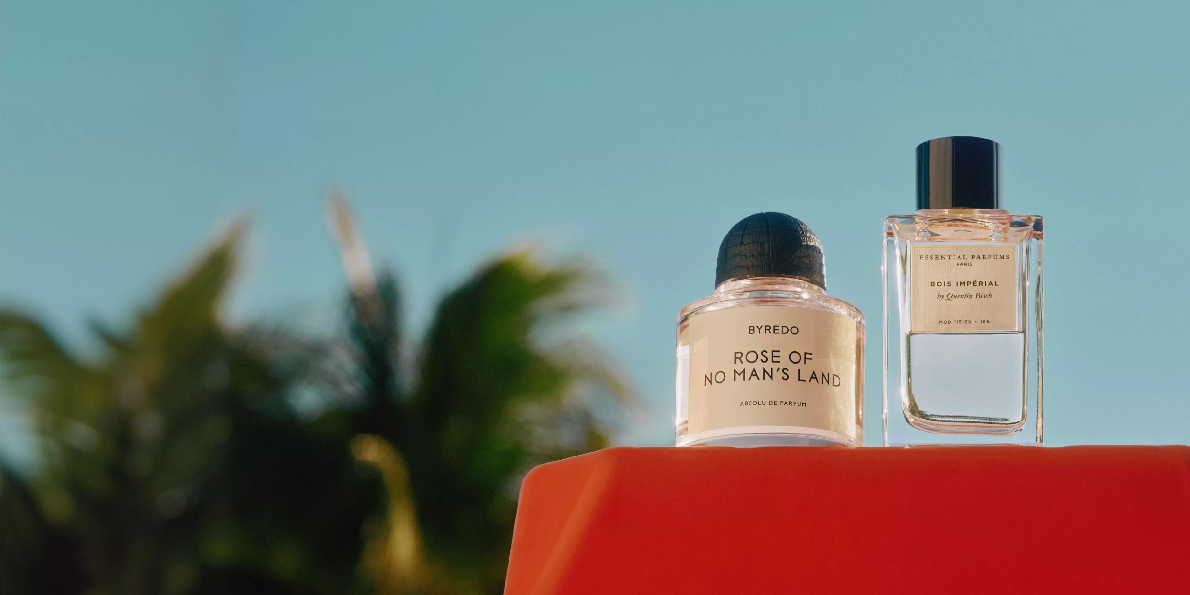 bottle of byredo - rose of no man's land perfume on a red table with sunny palm trees behind