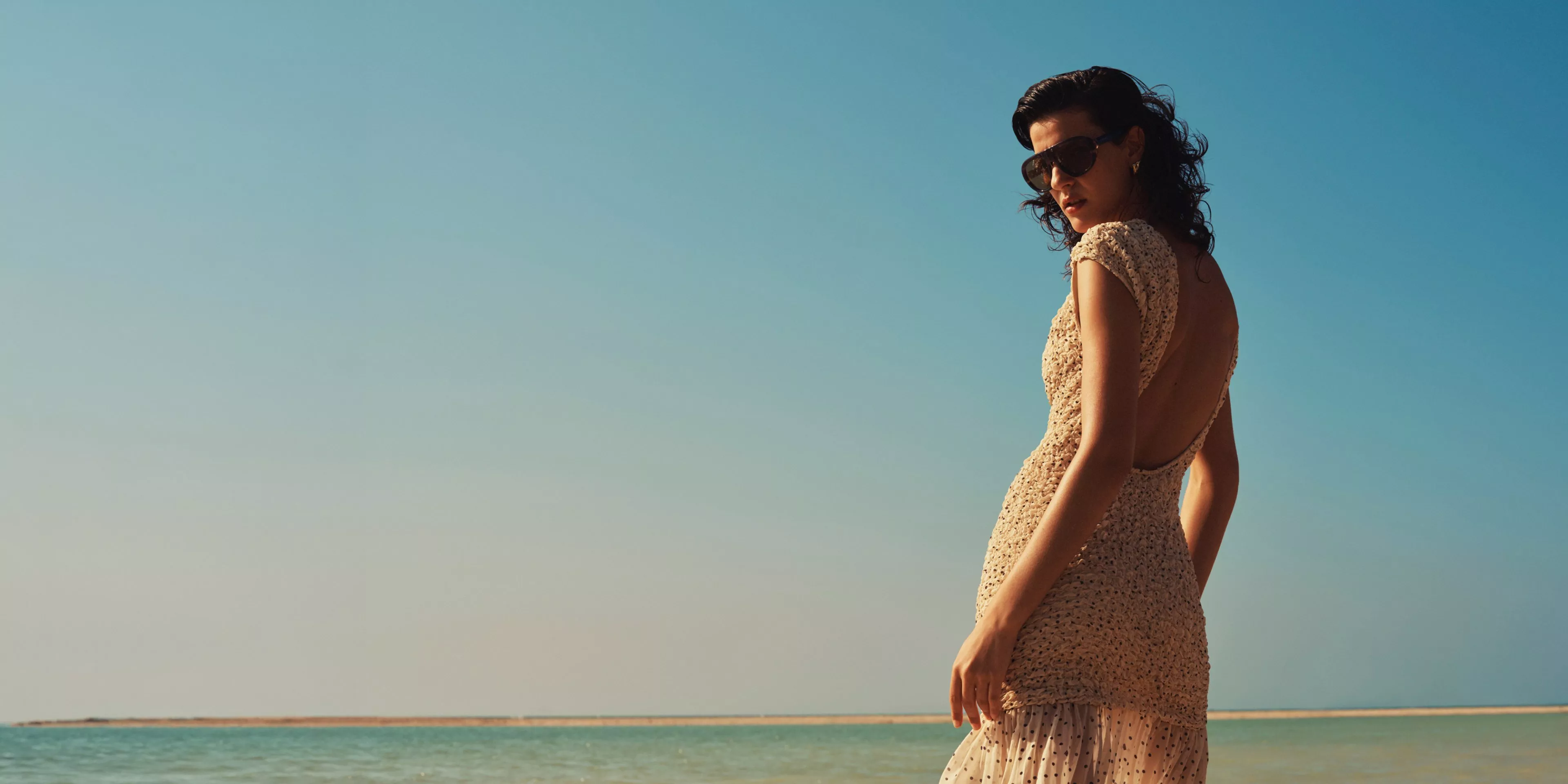 women in a dress with the sun hitting her face, with the sea behind her