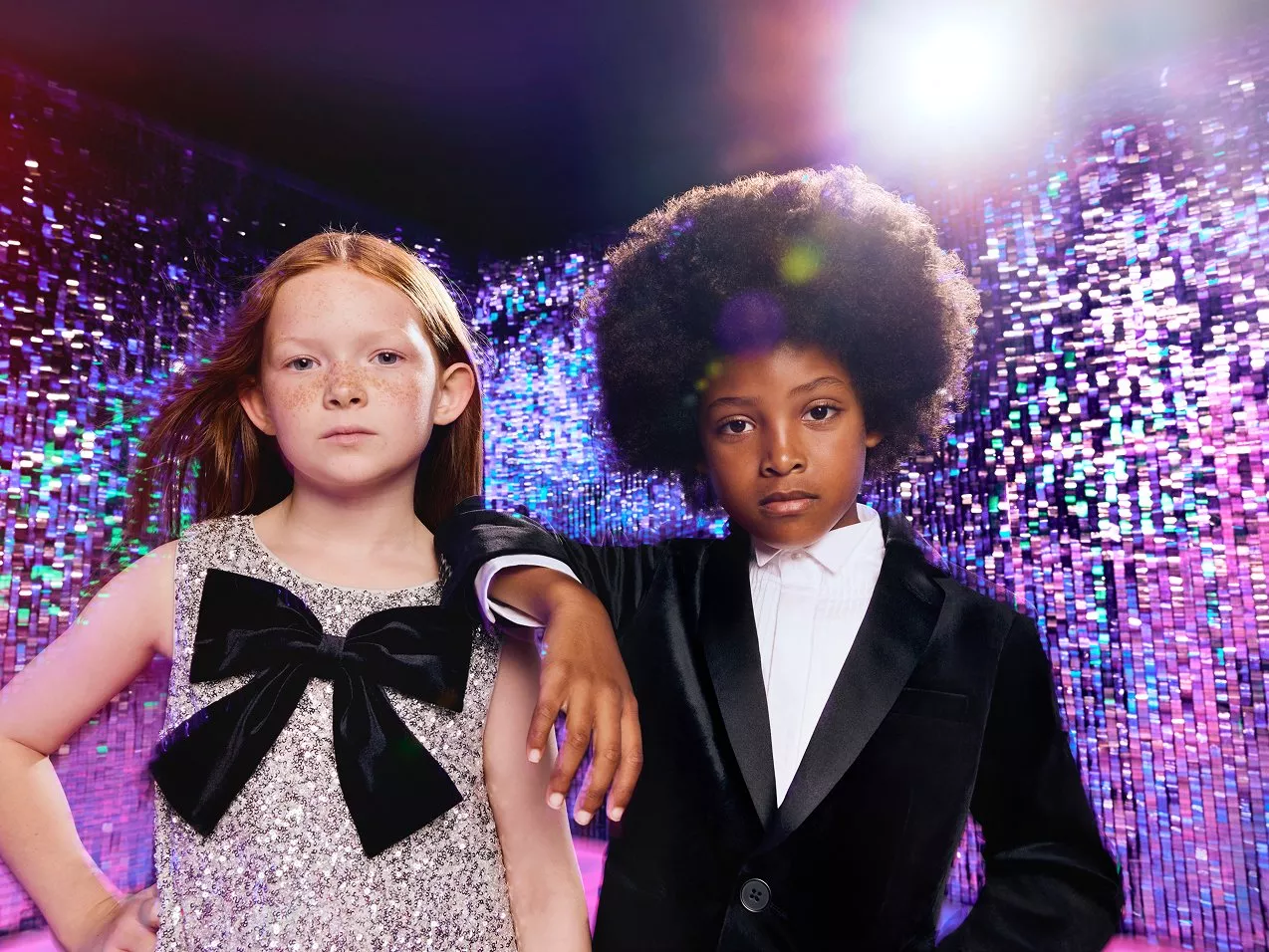 Girl in silver sequinned dress standing next to boy in velvet black suit and white shirt