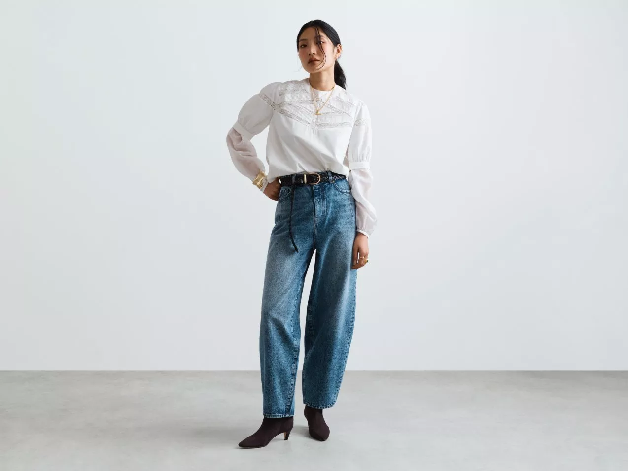 Full-length studio fashion photo of a woman in white lace blouse, wide-leg blue jeans, belt and ankle boots