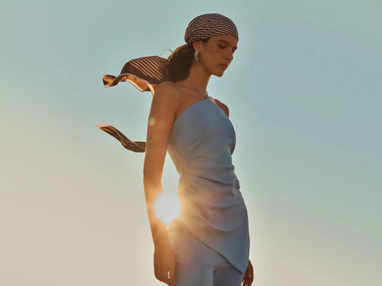 Fashion model in light blue evening dress with striped headscarf, standing at sunset against a clear sky.
