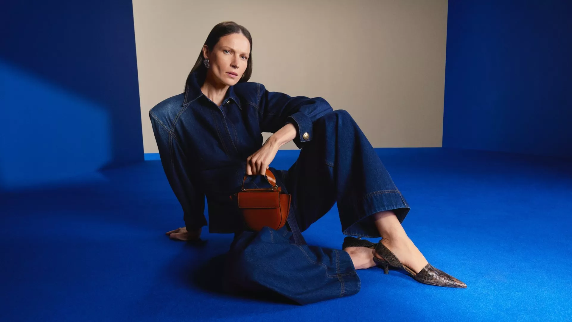 Fashion model in dark denim jumpsuit holding tan handbag, seated on blue studio set with neutral backdrop