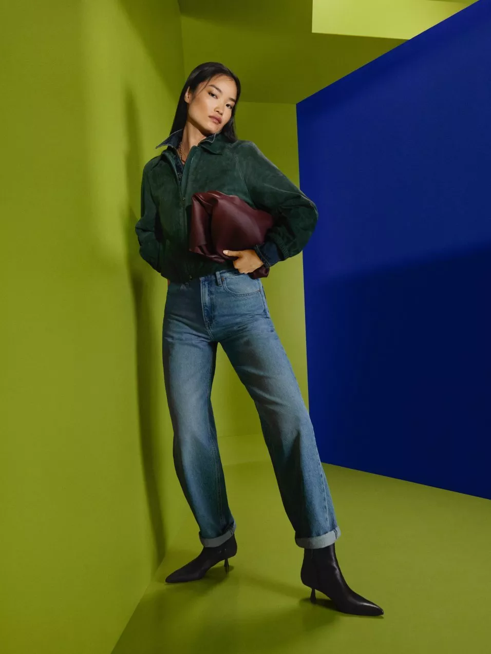 Fashion model in green suede jacket and blue jeans, holding burgundy clutch, studio backdrop in lime and blue