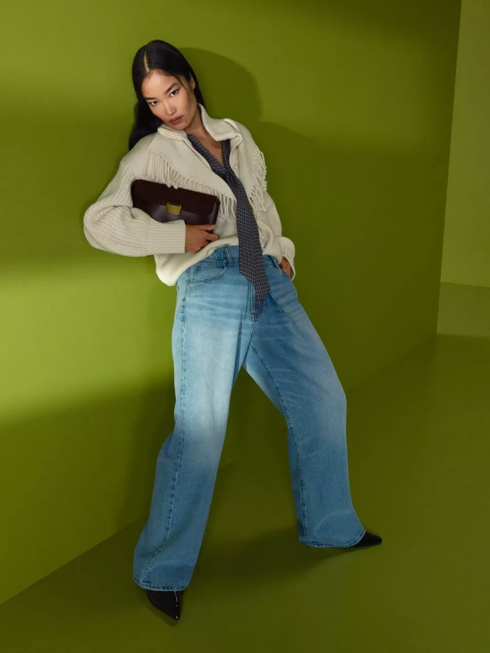 Fashion model in cream fringed cardigan, wide-leg jeans and heels holding a brown clutch against green backdrop