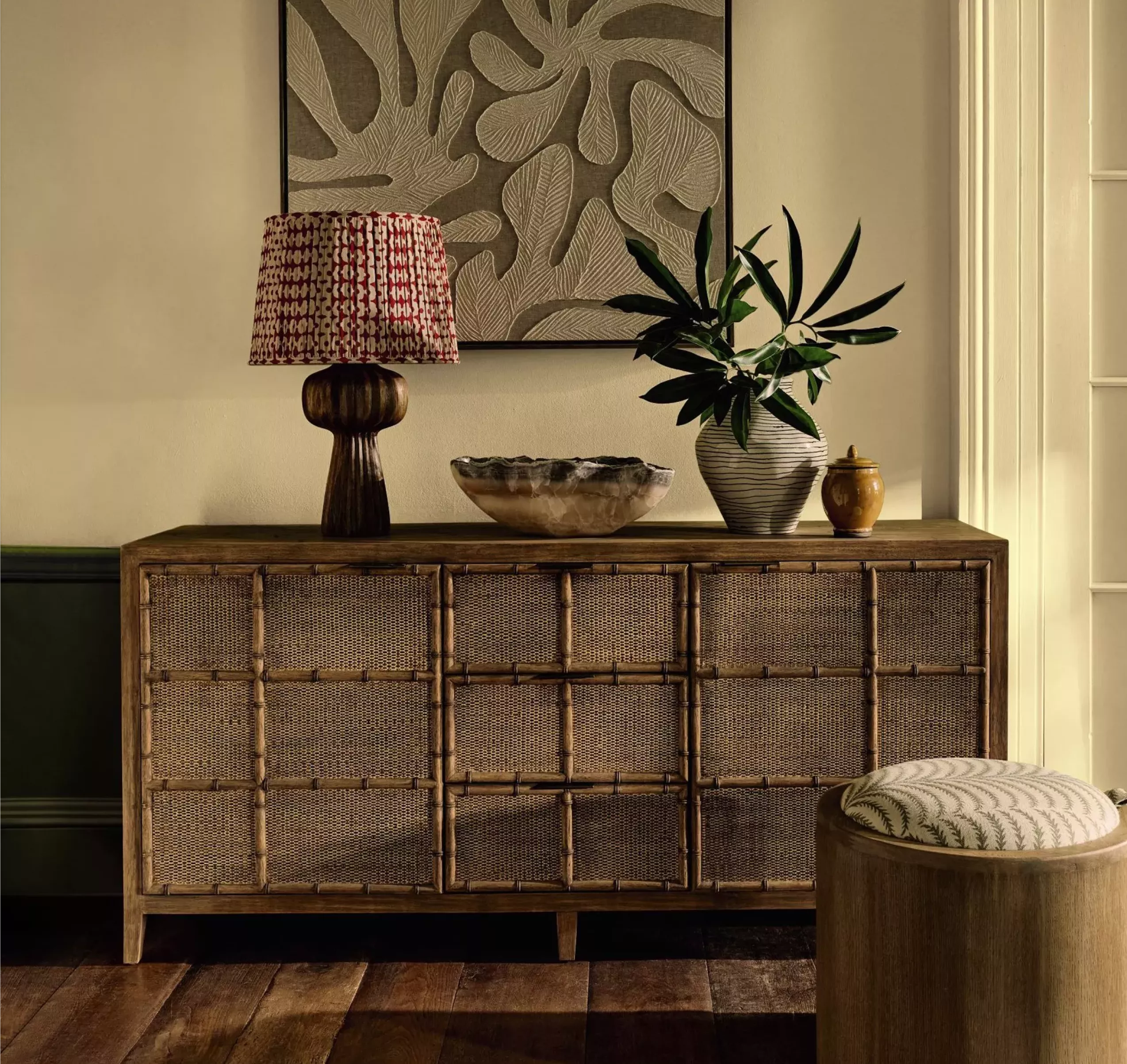 OKA Rattan sideboard with table lamp, ceramic bowl and houseplant beneath abstract wall art in stylish living room interior