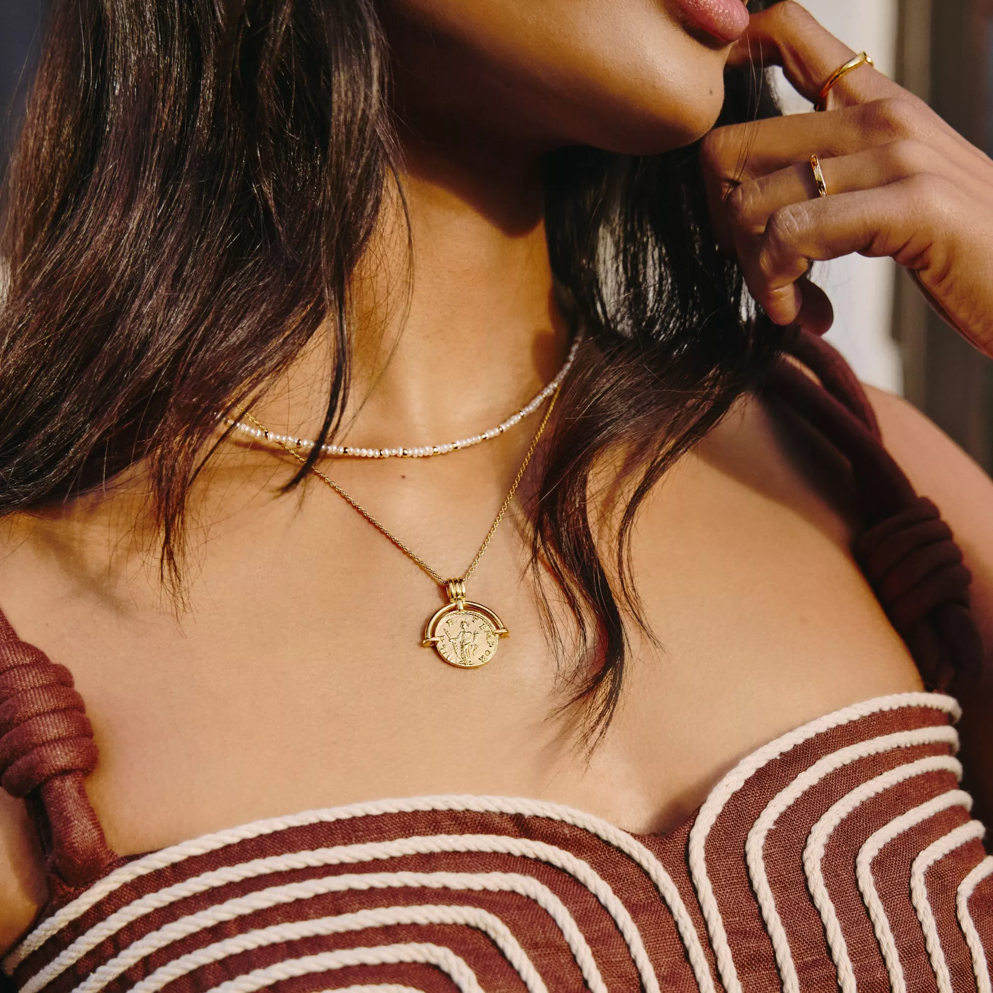 Close-up of woman wearing layered gold coin pendant necklace and pearl choker, with gold rings in sunlight