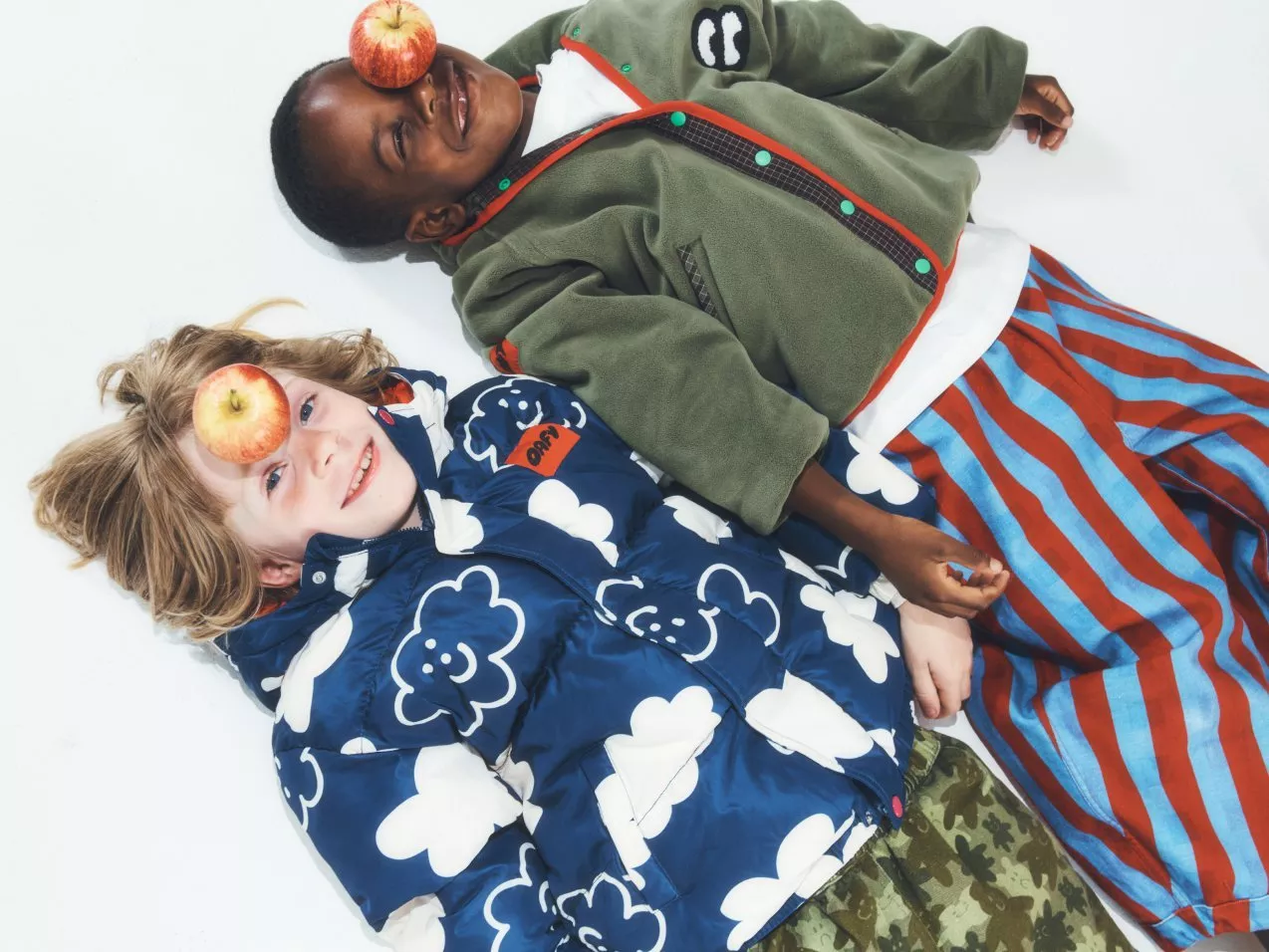 Two children lying on a white studio floor with apples on their faces, wearing colourful winter jackets and trousers