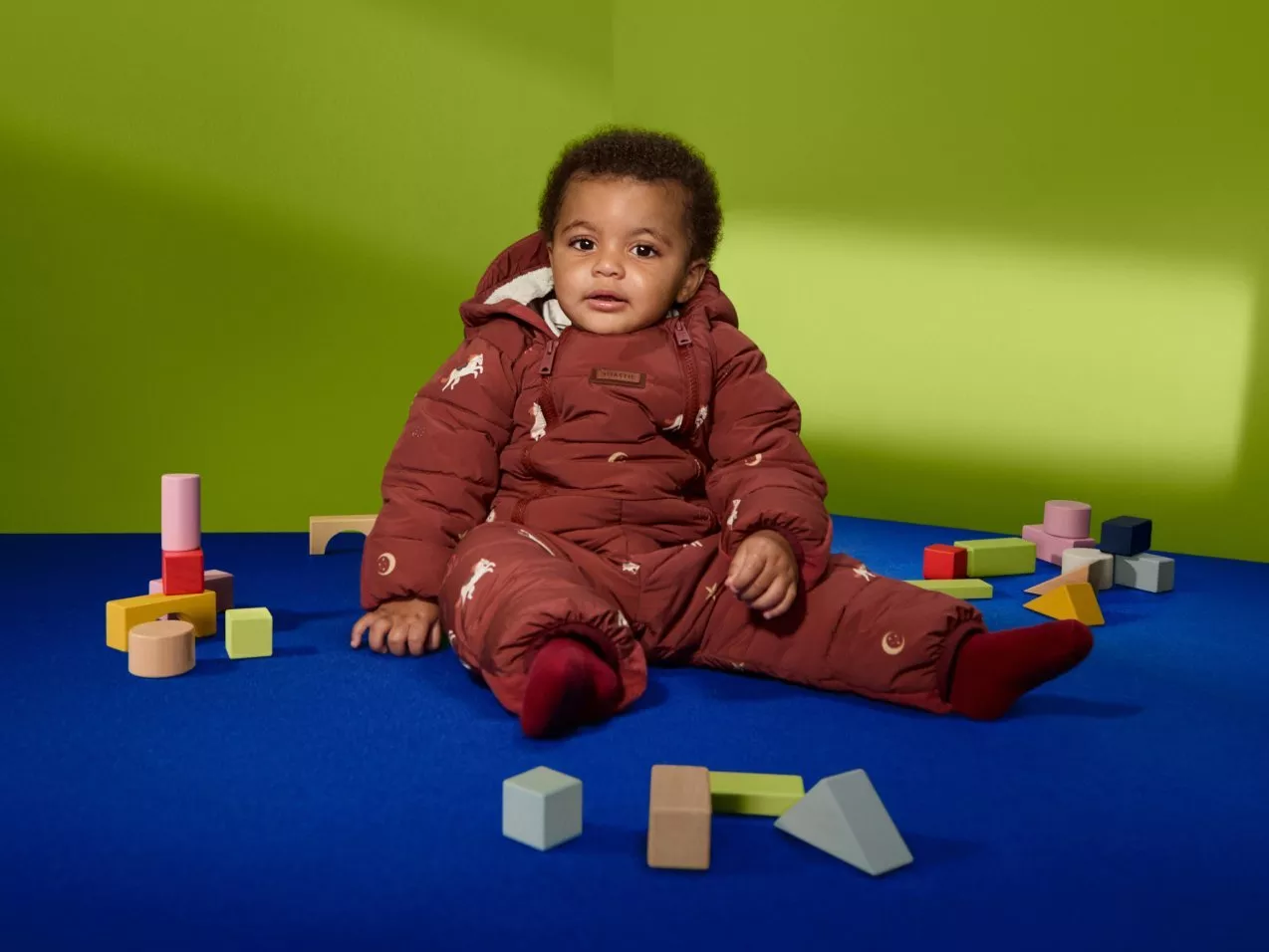 Toddler wearing a brown padded snowsuit sits on blue floor with colourful wooden blocks, lime green backdrop
