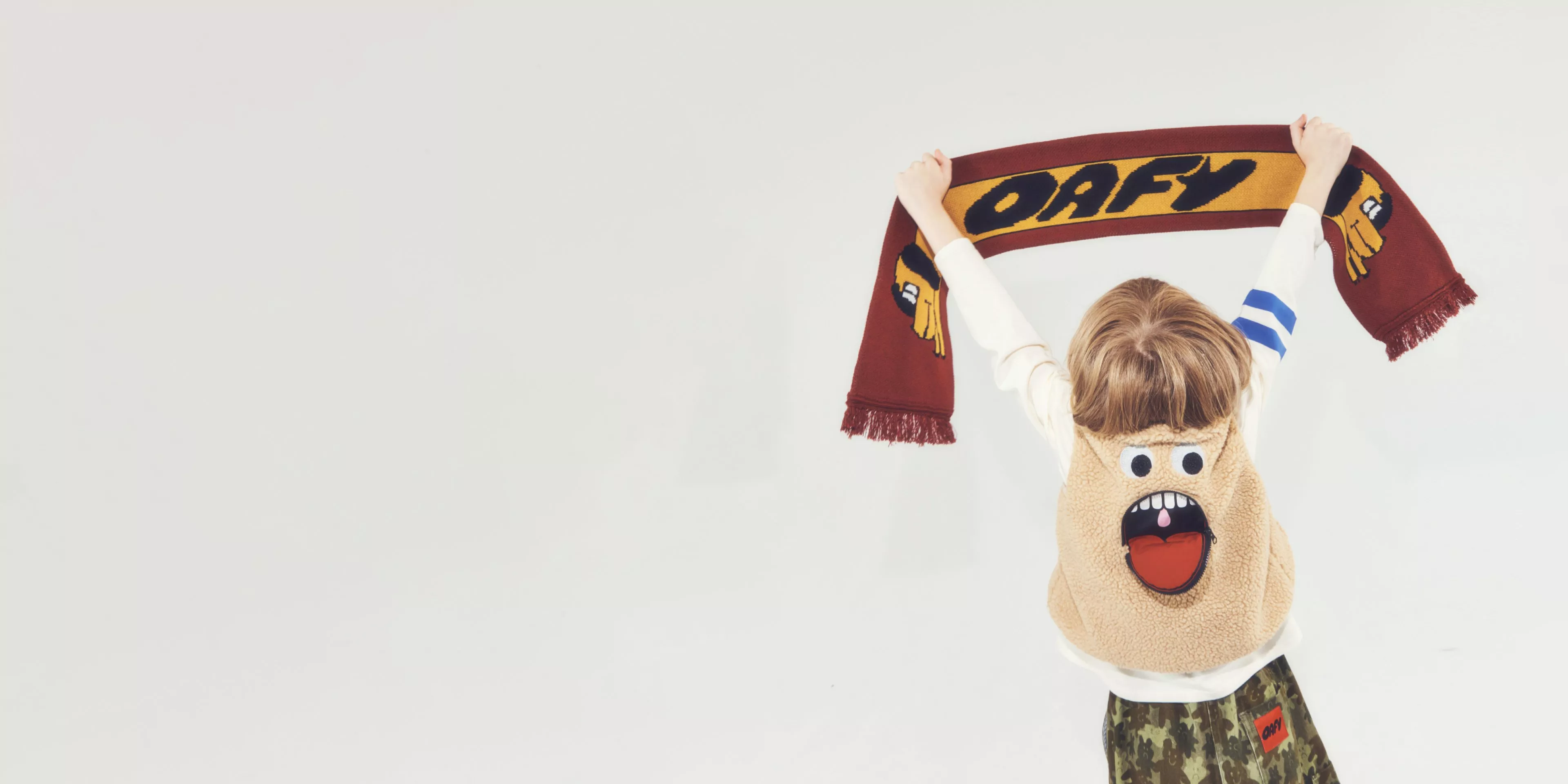 Child holding an OAFY scarf overhead, wearing a cartoon face fleece gilet and camo trousers on a plain backdrop