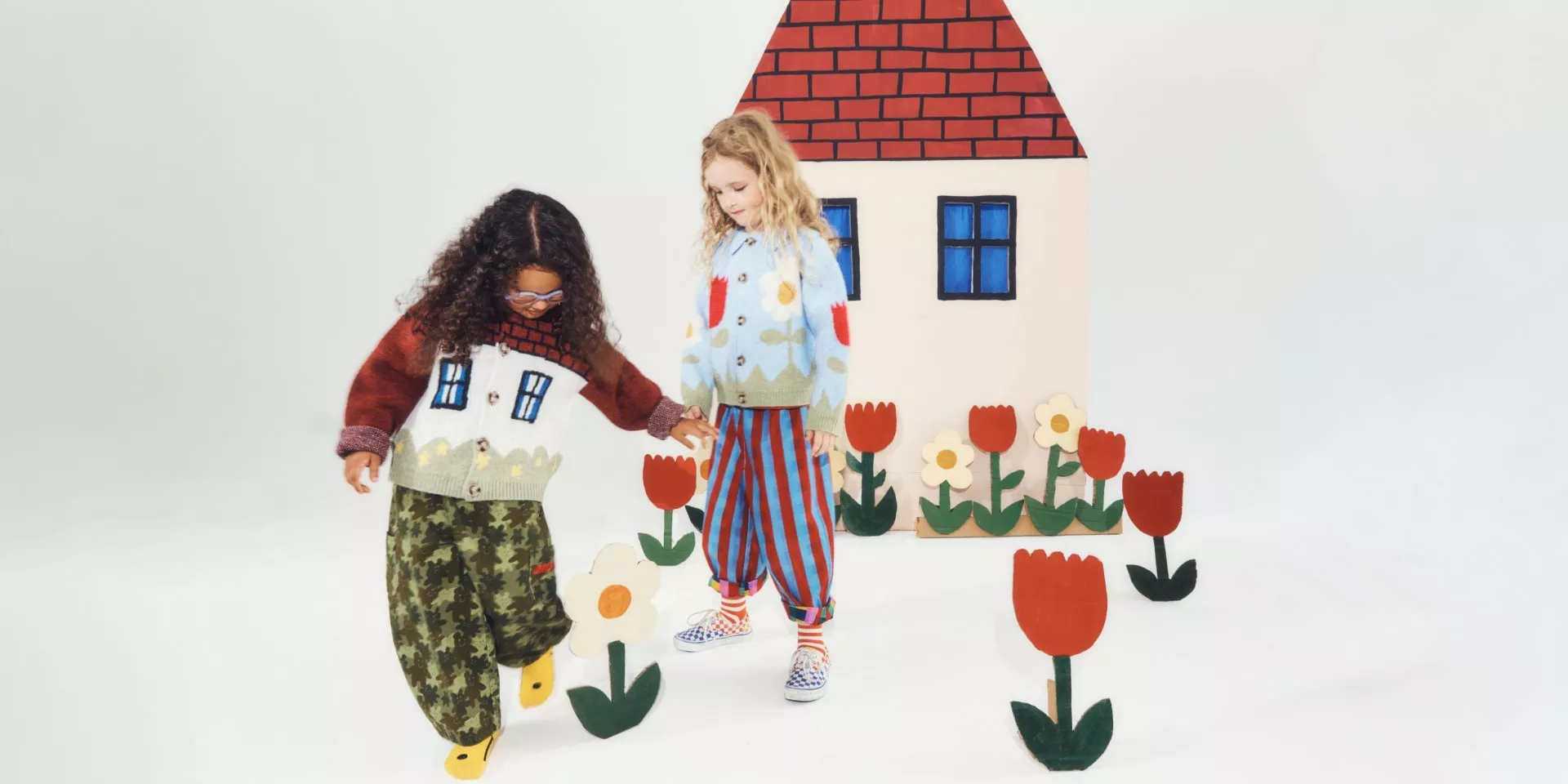 Two children in colourful knit cardigans and trousers pose by a playful house set with tulip props.