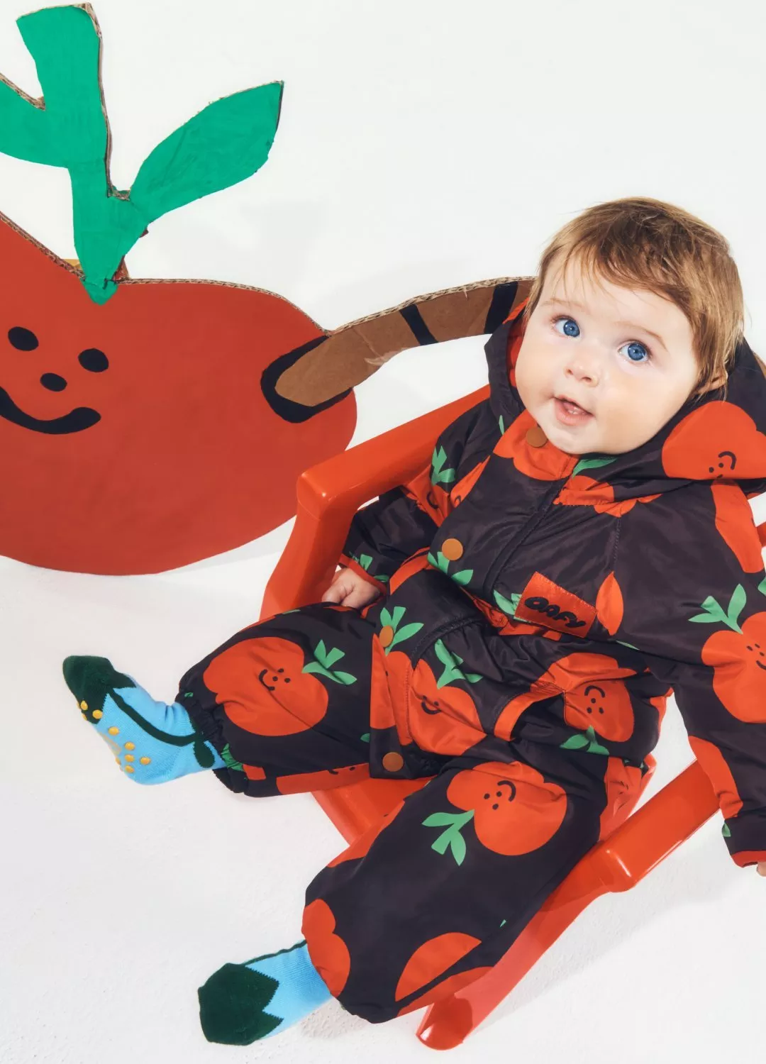 Baby in apple-print snowsuit sitting on red chair beside cardboard apple decoration, studio photo