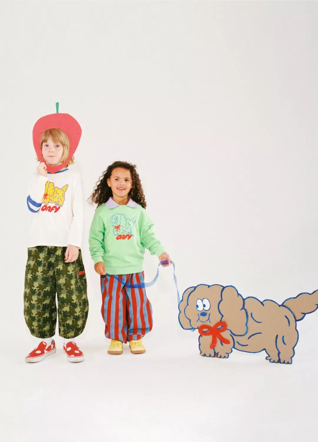 Two children in playful outfits, one wearing an apple headpiece, holding a lead to a cartoon dog cut-out