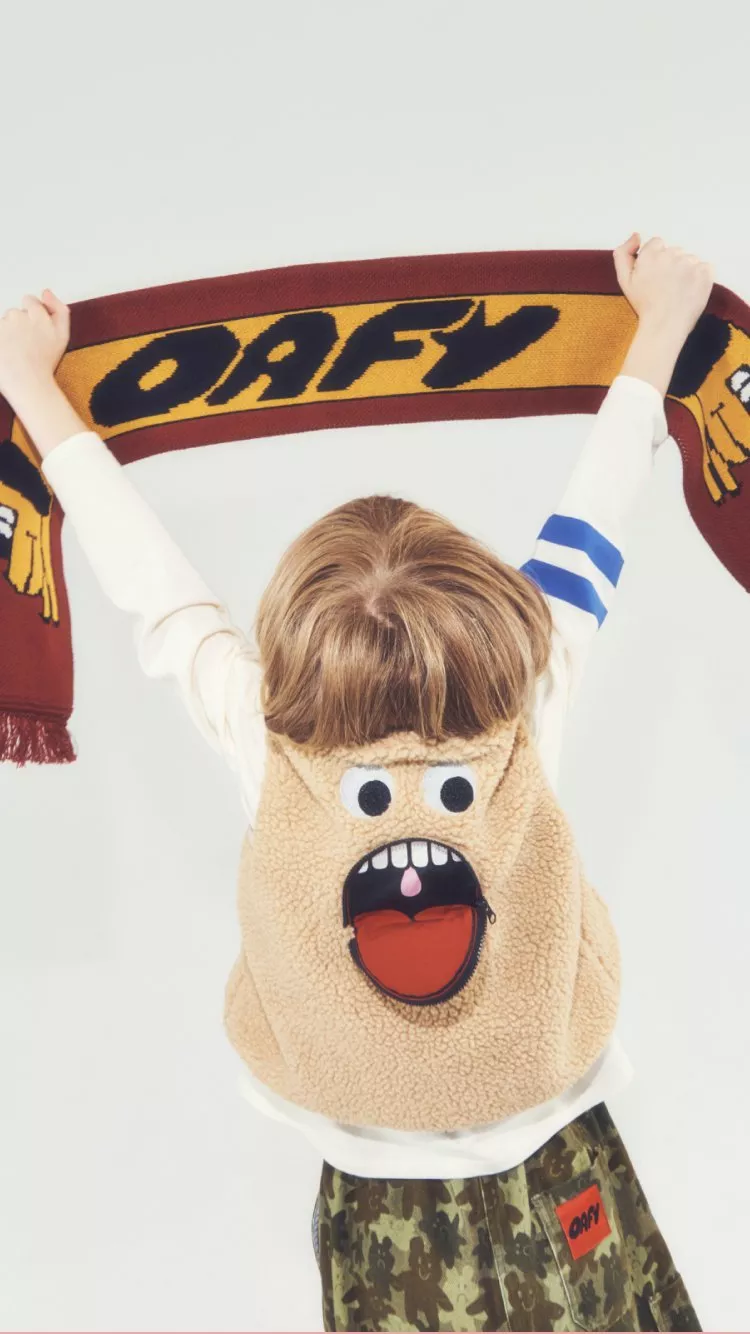 Child holding OAFY scarf overhead, wearing fluffy vest with cartoon face and camouflage trousers on white background