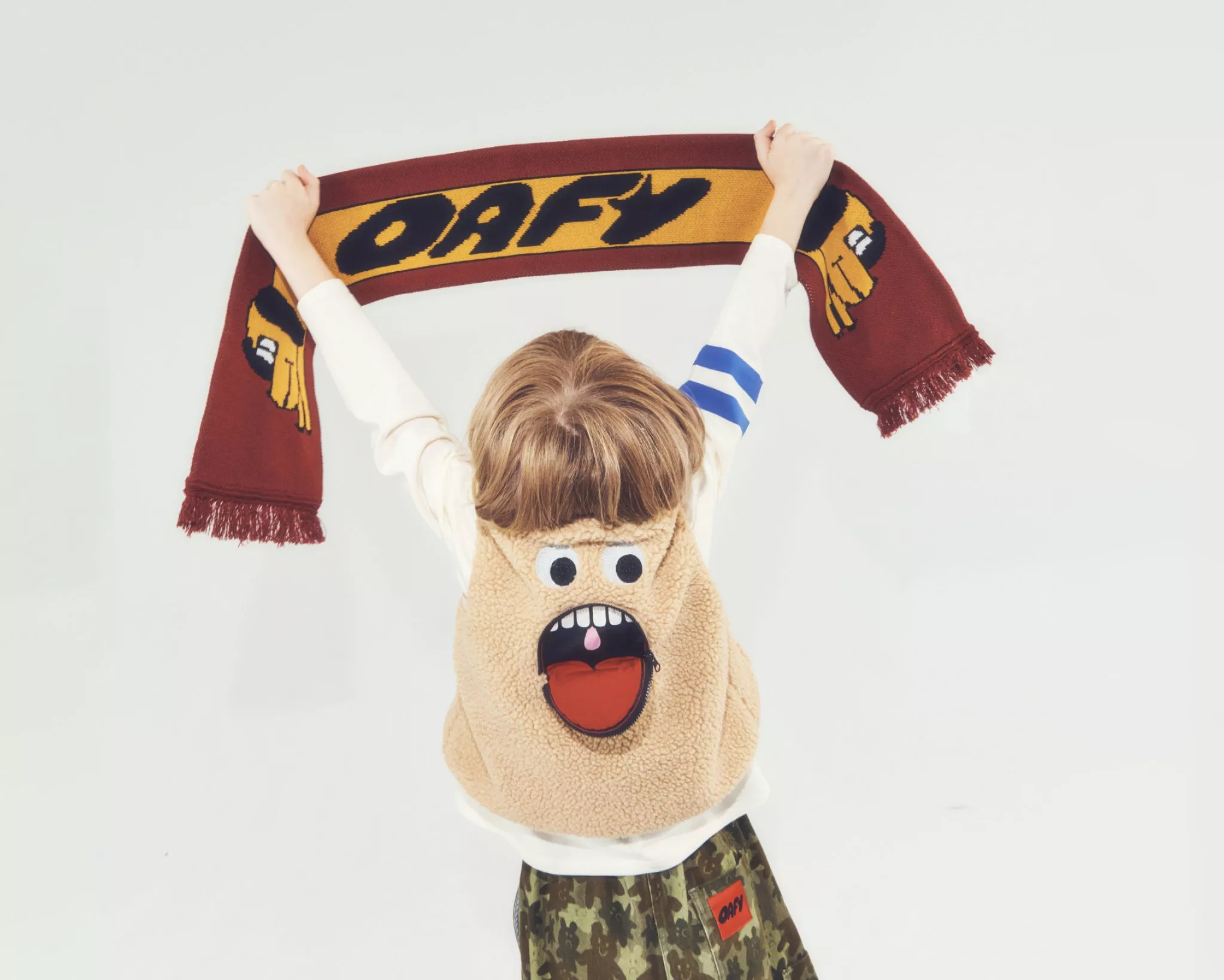  Child wearing Oafy by Lazy Oaf fleecy gilet with cartoon face on the reverse, holding an Oafy striped scarf in the air