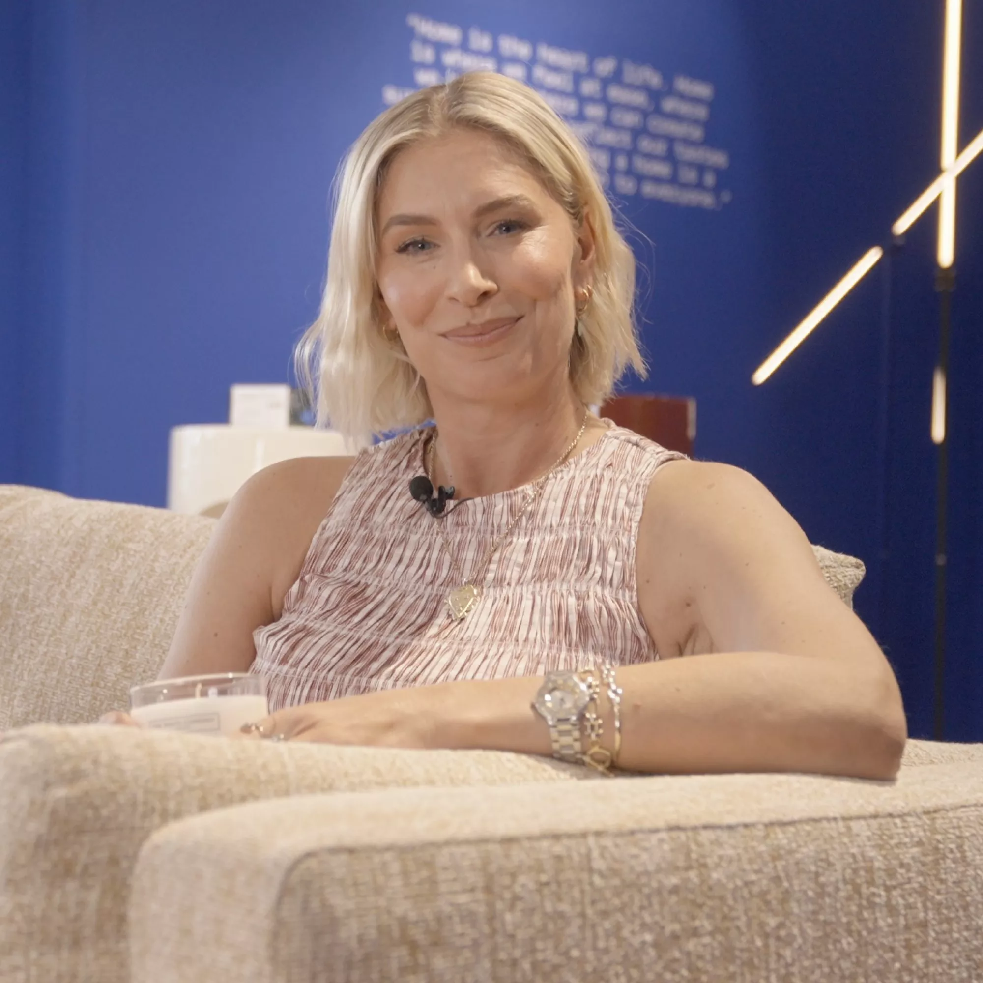 Smiling blonde woman seated on a beige sofa in a modern lounge, wearing a lapel mic and jewellery.