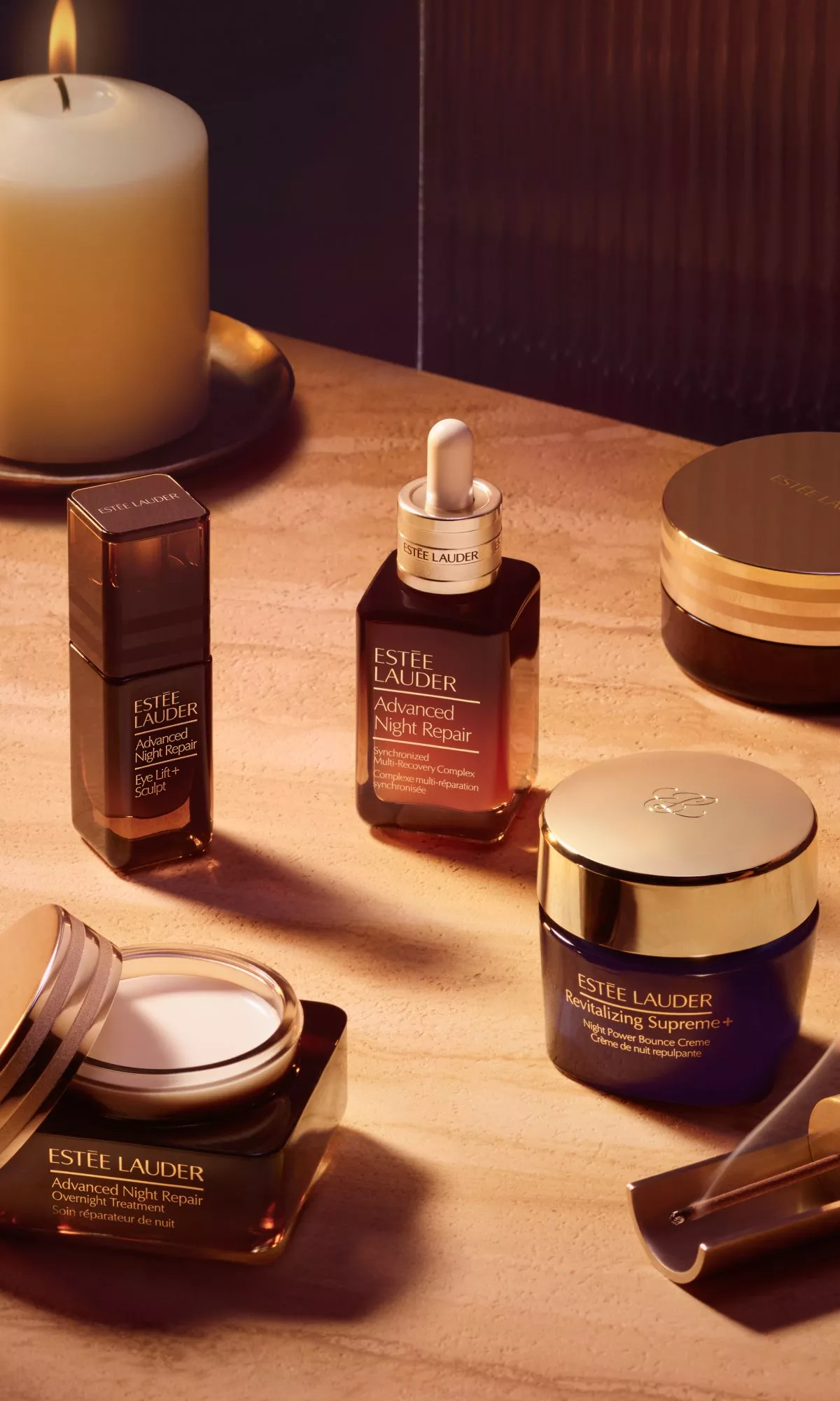 A collection of Estée Lauder products, including Advanced Night Repair serum and Revitalizing Supreme+ cream, arranged on a textured surface under warm, golden light.