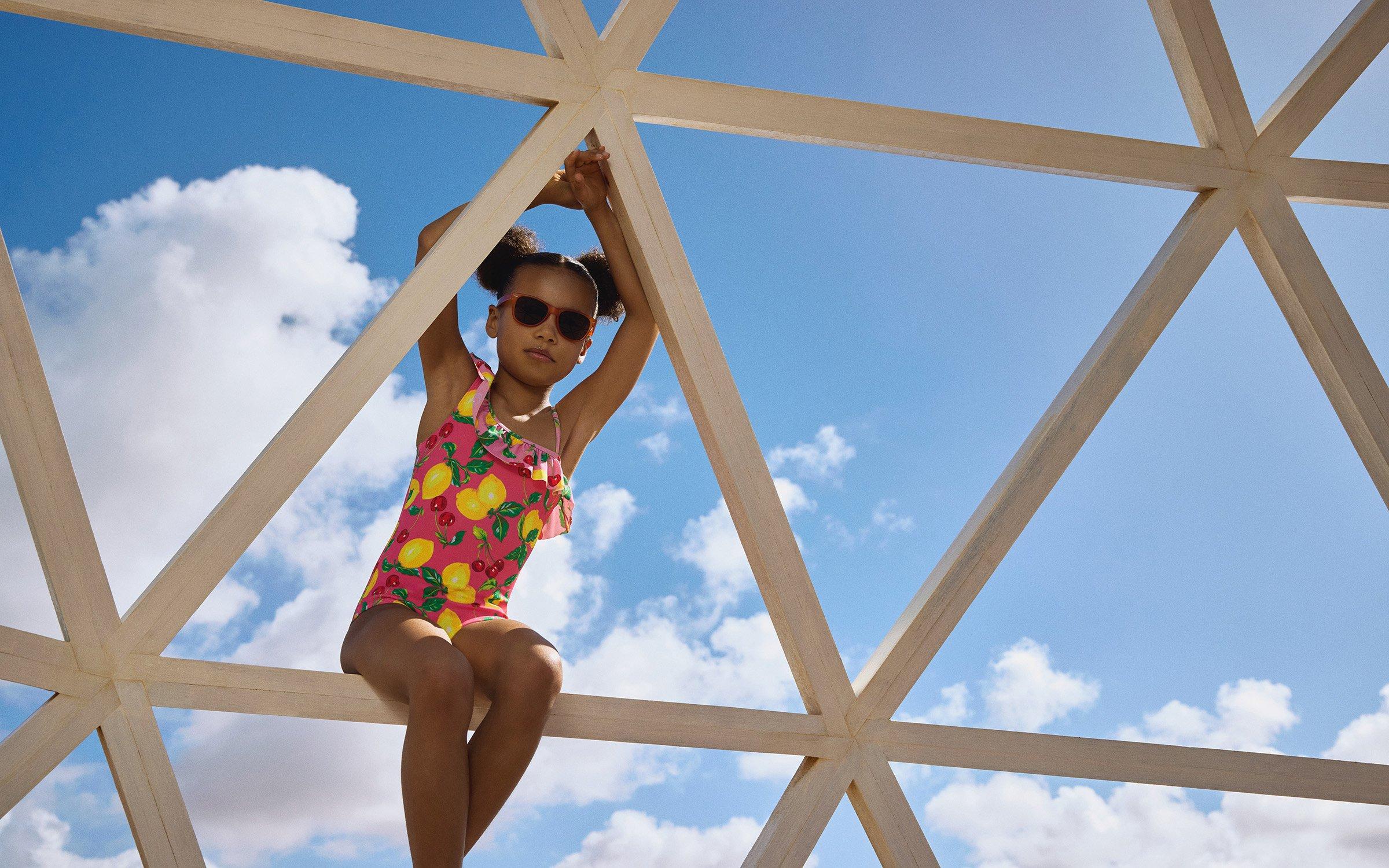 Kid in swimming costume and sunglasses, sitting on a wooden frame against a sky backdrop.
