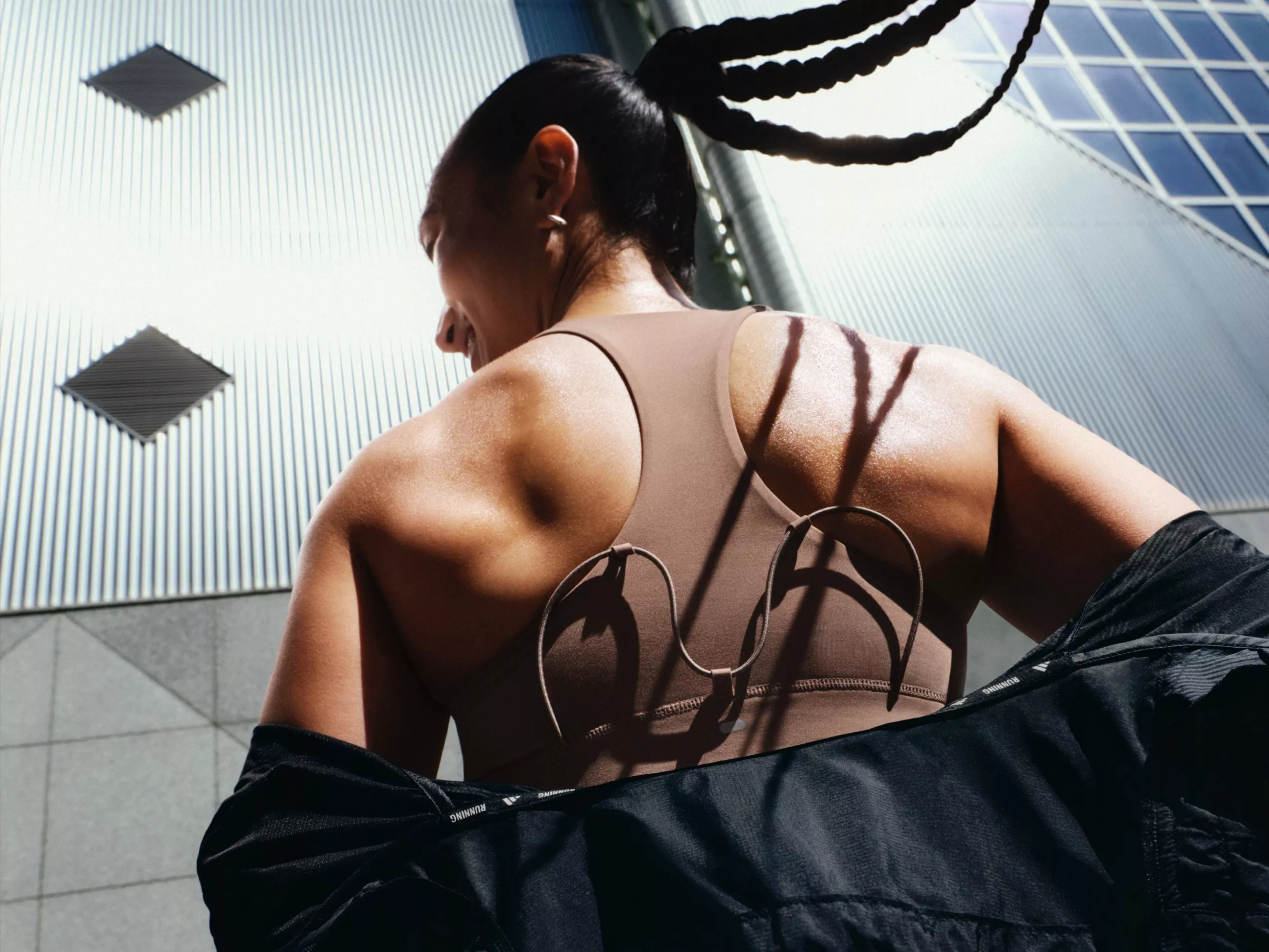 Back view of athletic woman in sports bra and jacket outdoors, with braided ponytail near modern buildings