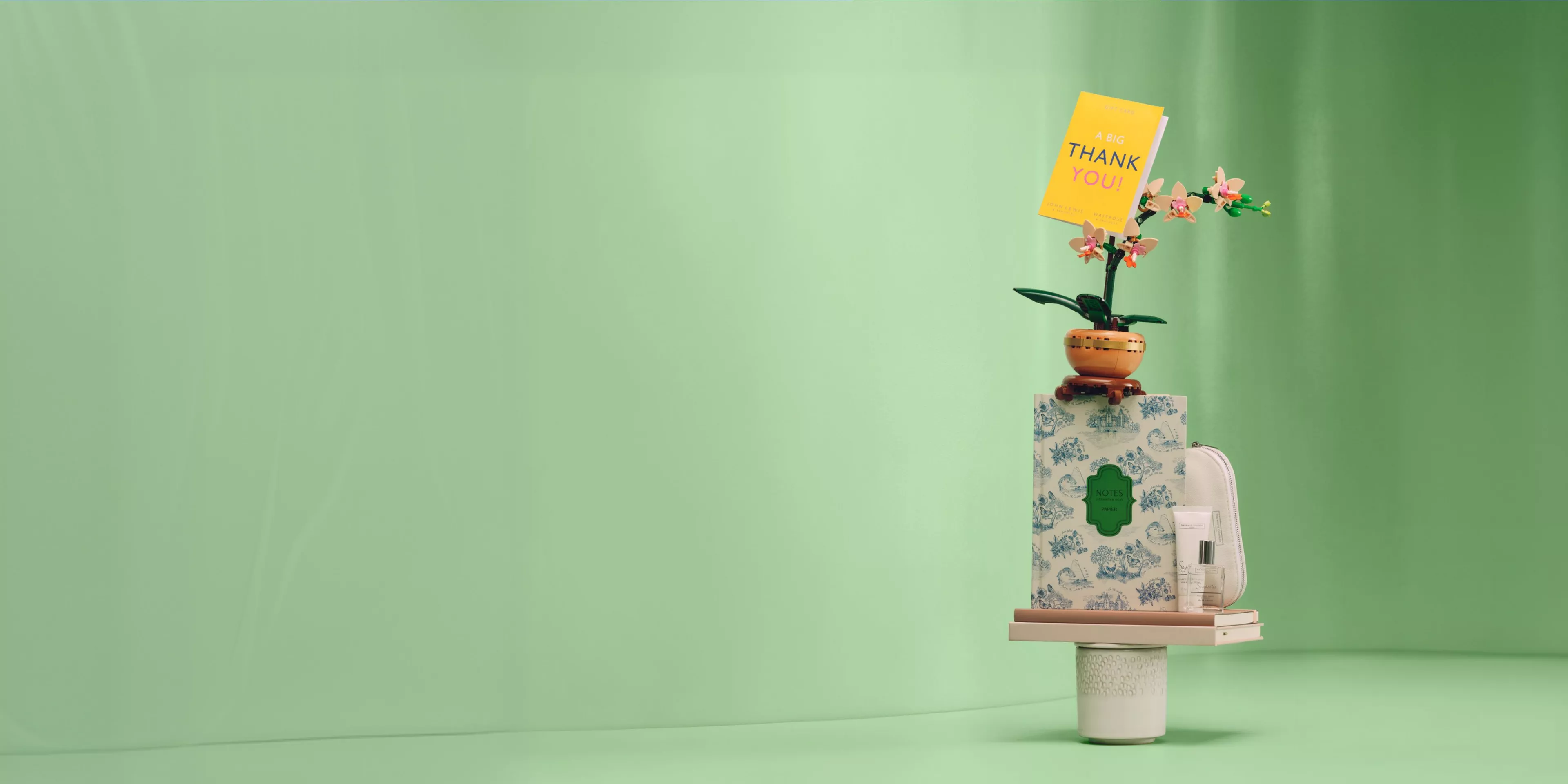 Gift hamper with thank you card, orchid plant, floral notebook, perfume and toiletry bag on green backdrop