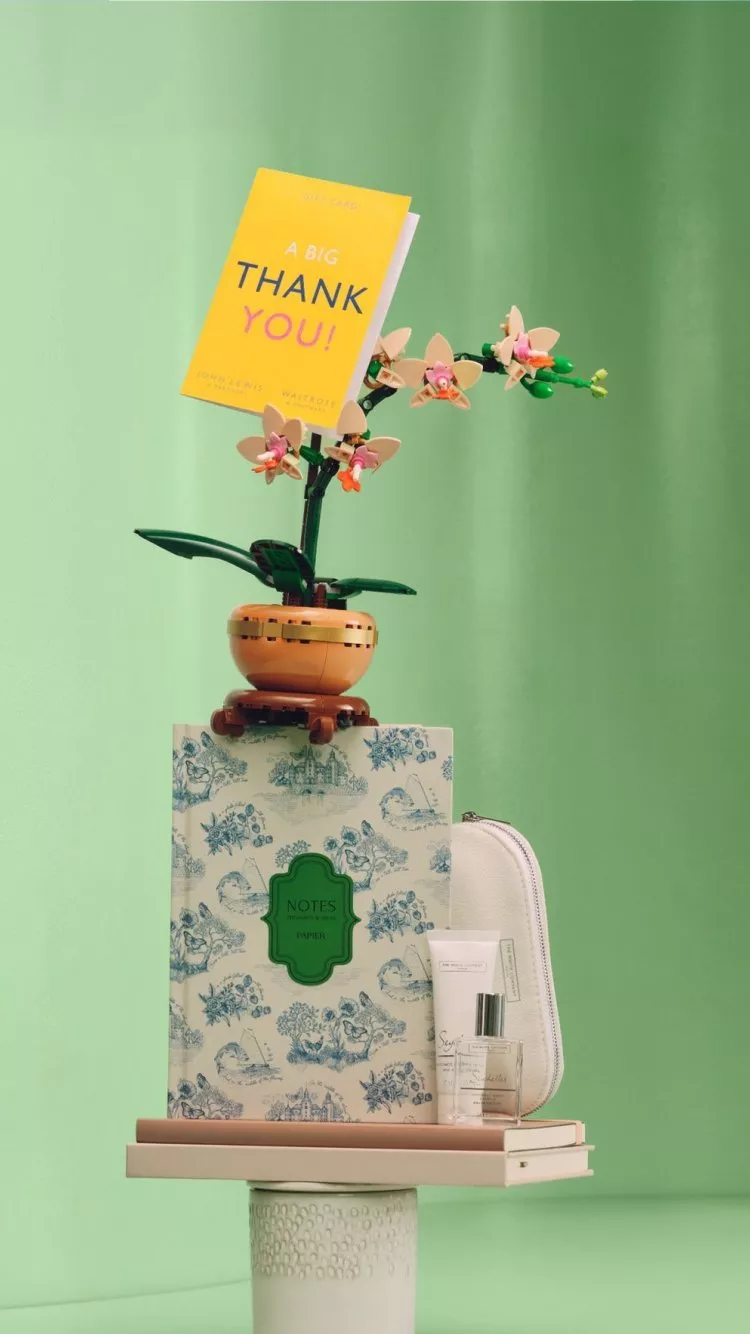 Gift hamper with thank you card, orchid plant, floral notebook, perfume and toiletry bag on green backdrop