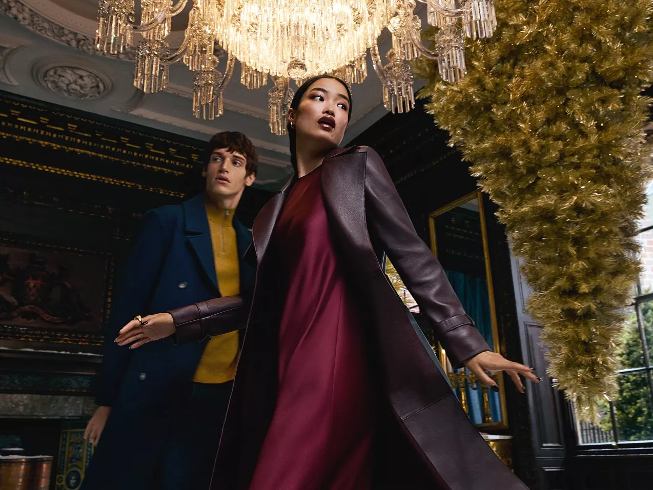 Man in blue jacket and yellow sweater, and woman in brown leather coat and red dress under chandelier