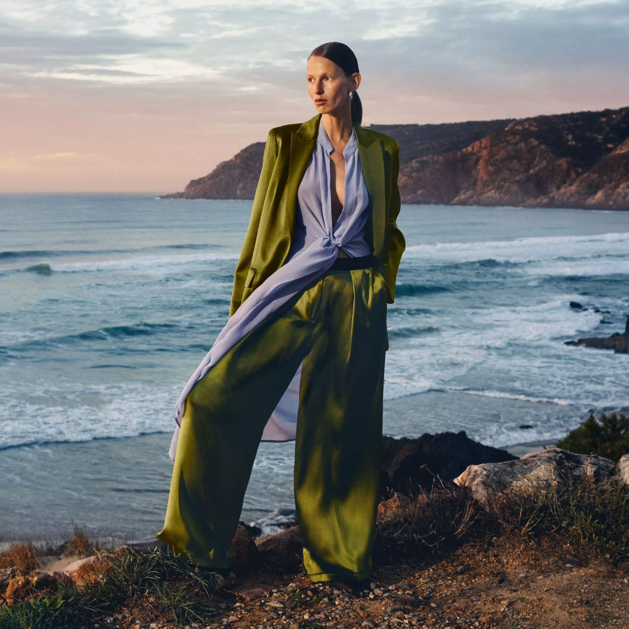 Fashion model in green suit and blue blouse posing on rocky coastline at sunset, waves and cliffs behind