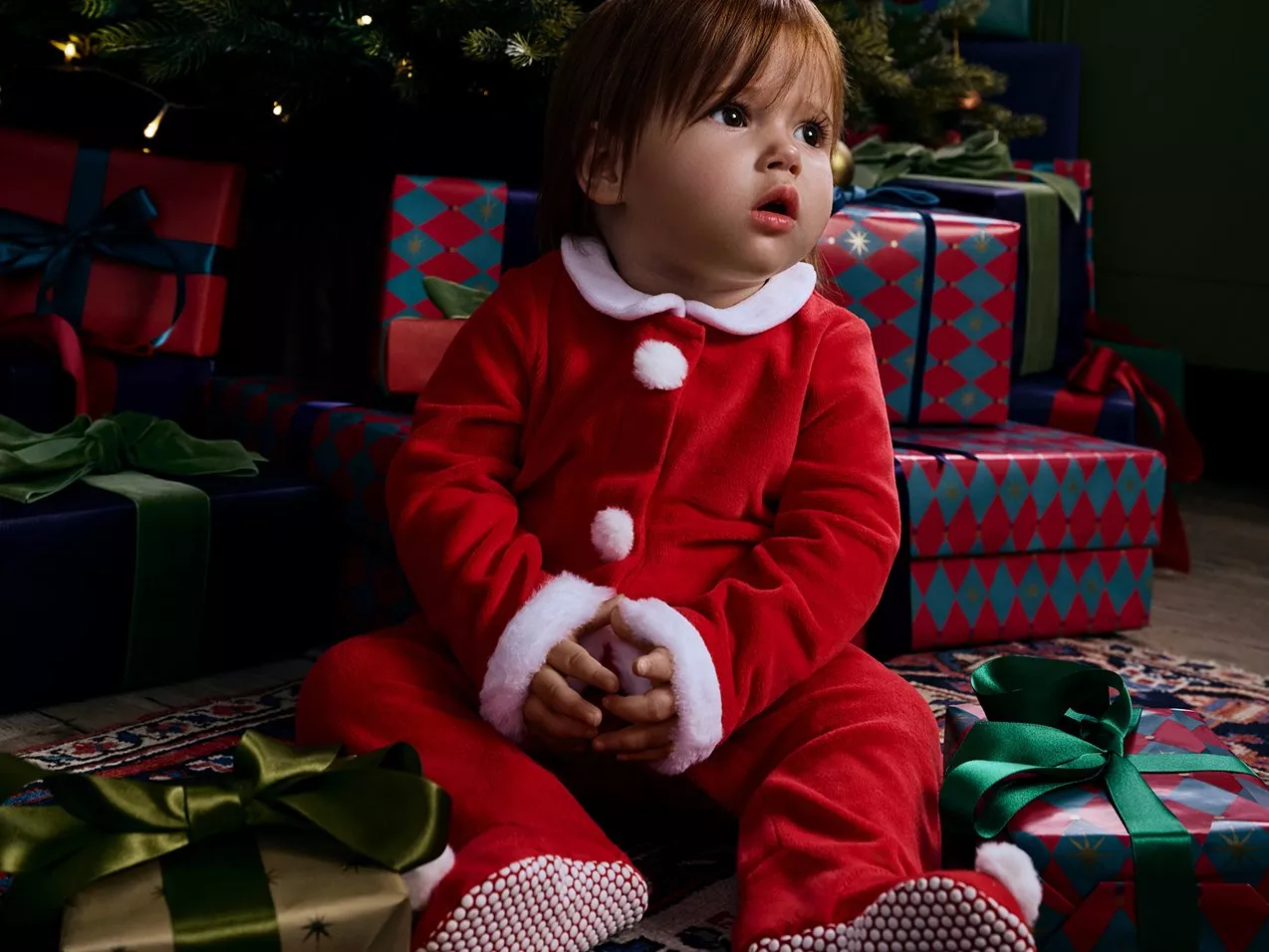 Toddler sitting in front of Christmas tree and gifts in a red Christmas onsie