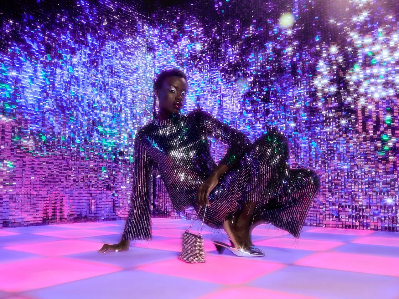 A person wearing a shimmering, silver sequined outfit squats on a glowing pink and blue checkered dance floor in front of a sparkling disco-ball-style background.