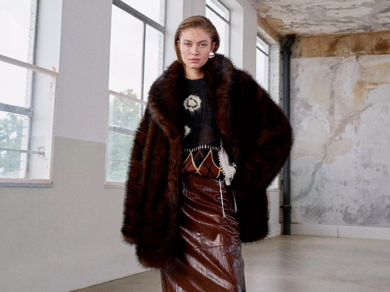 A woman in a brown fur coat and patterned sweater, shiny burgundy leather effect skirt posing in an industrial-style room