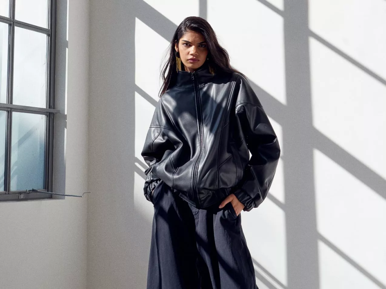 Woman wearing a black leather jacket and wide trousers, standing indoors near a window.