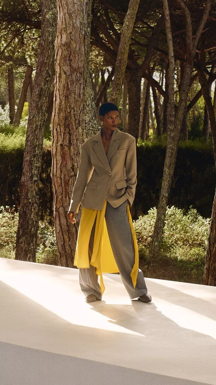 A person in a beige suit and yellow skirt stands outdoors amongst trees and greenery in soft sunlight.