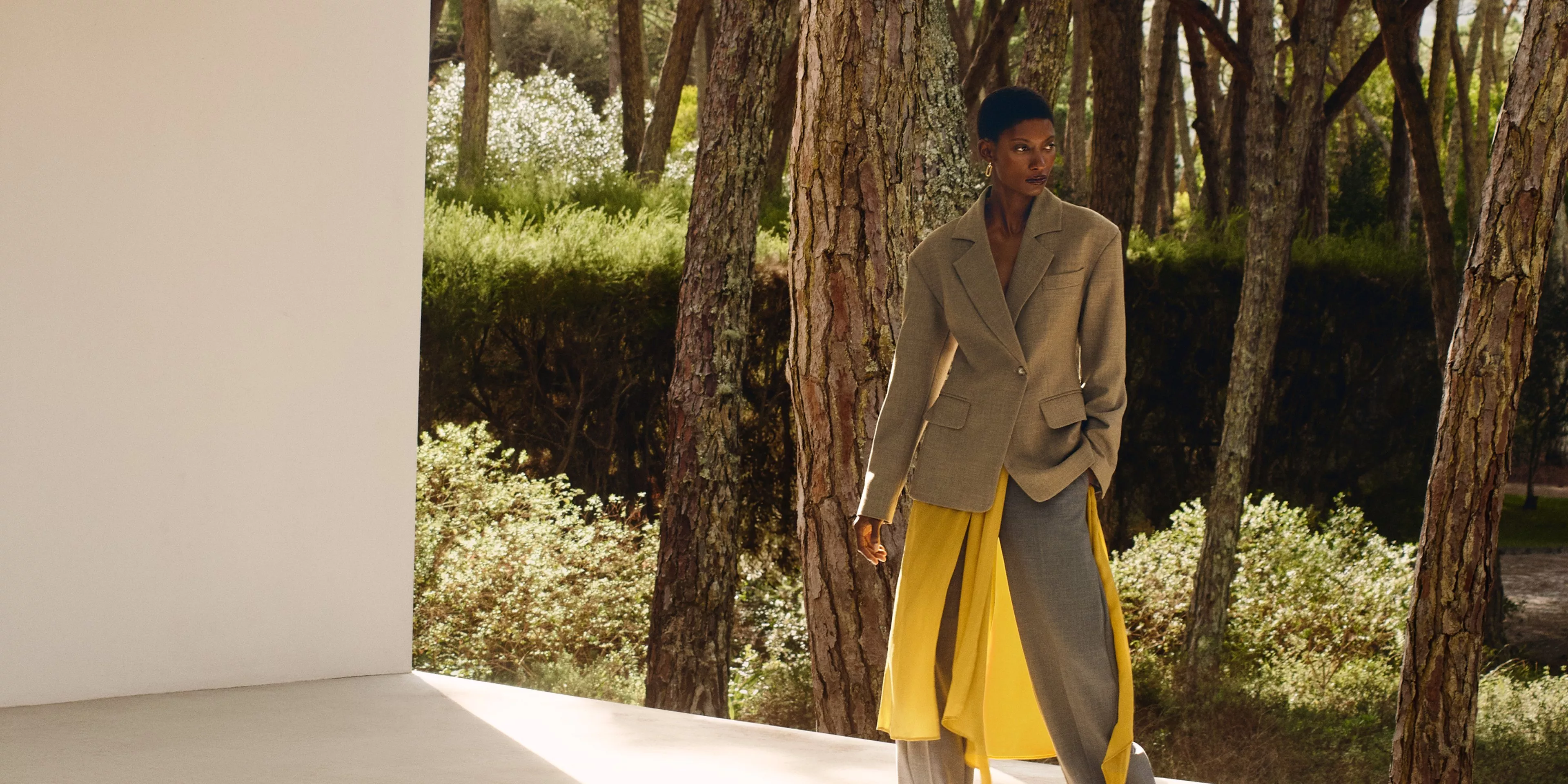 Fashion model in a stylish beige blazer and yellow skirt stands in a serene wooded area.