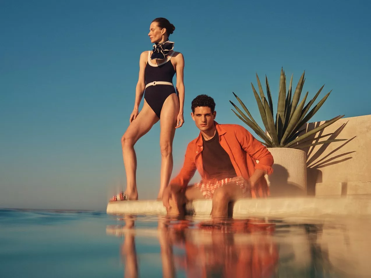 Stylish couple by a pool at sunset, woman in black swimsuit and scarf, man in orange shirt, agave backdrop